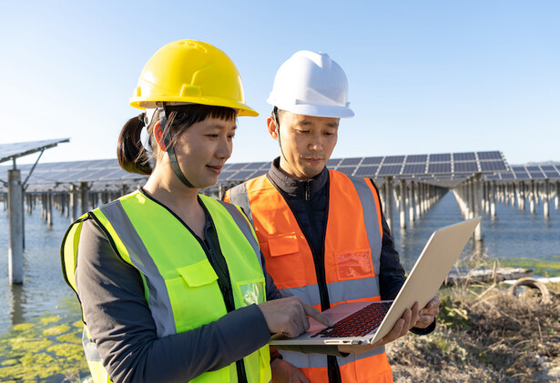 Promo image for Carbon reduction, of two young workers with hard hats, hi-vis jackets looking at a laptop with solar panel field in background